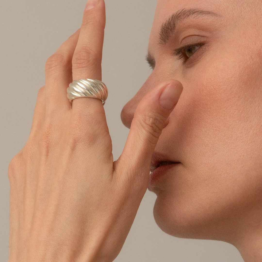 A close-up lifestyle shot of a model wearing a wide, textured silver ring on her finger, touching her nose with the other hand against a soft beige background.