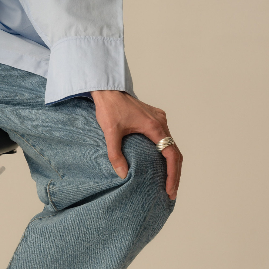 A close-up lifestyle shot of a person wearing a wide, textured silver ring on their hand, dressed in a light blue shirt and blue jeans against a solid beige background.