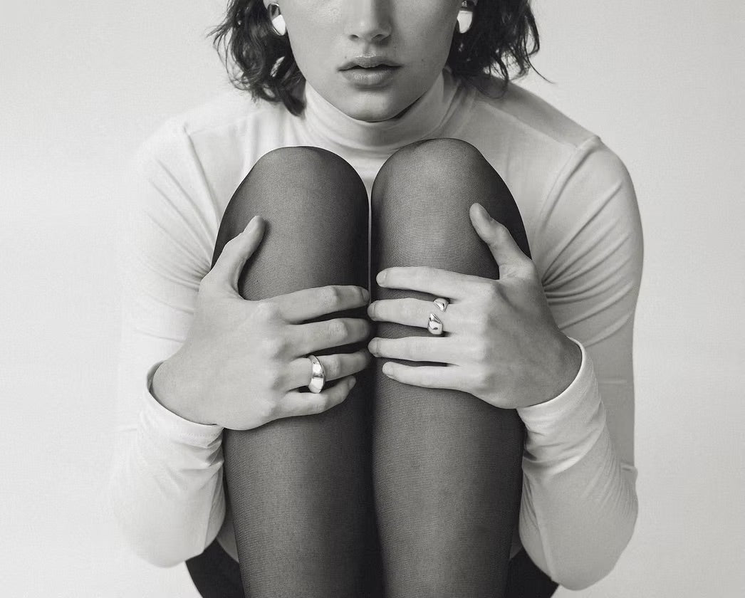 A black and white close-up of a person sitting with knees drawn up, wearing a fitted long-sleeve top, sheer black tights, and several rings on their fingers against a plain light background.