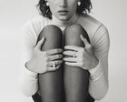 A black and white close-up of a person sitting with knees drawn up, wearing a fitted long-sleeve top, sheer black tights, and several rings on their fingers against a plain light background.