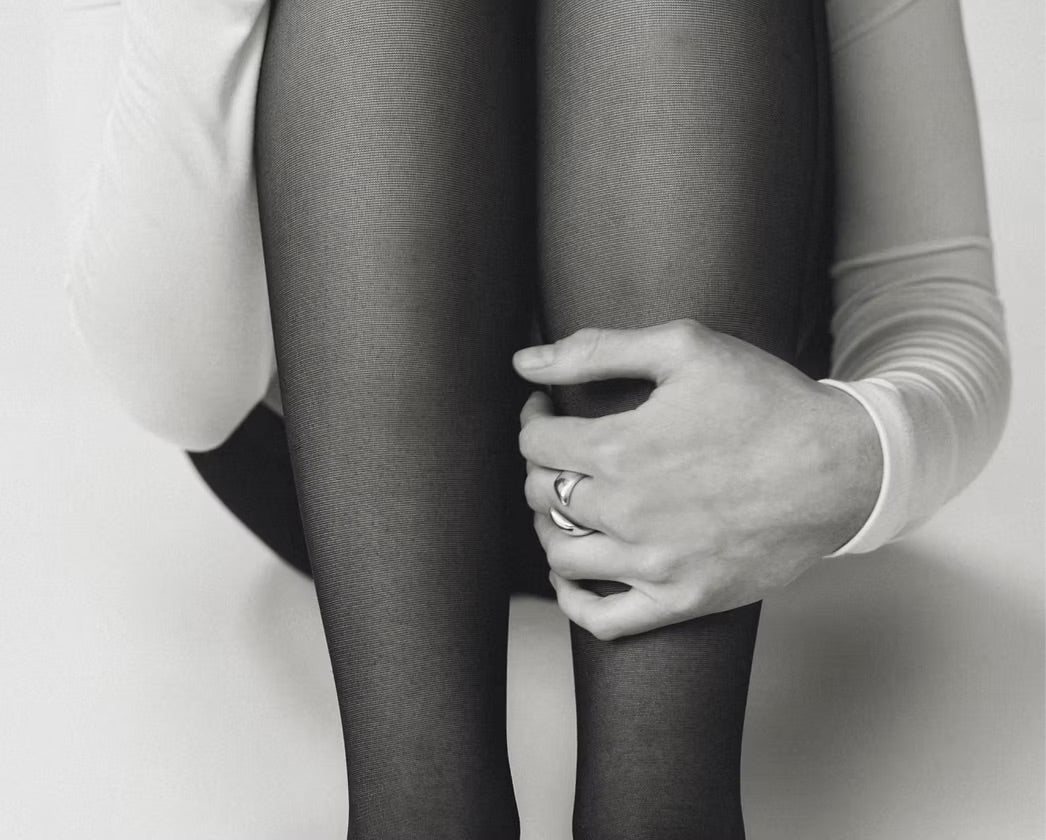 Black and white close-up of a person wearing sheer black tights, with one hand resting on the knee. The person is wearing a white long-sleeve top and a ring on one finger.