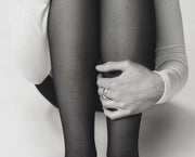 Black and white close-up of a person wearing sheer black tights, with one hand resting on the knee. The person is wearing a white long-sleeve top and a ring on one finger.