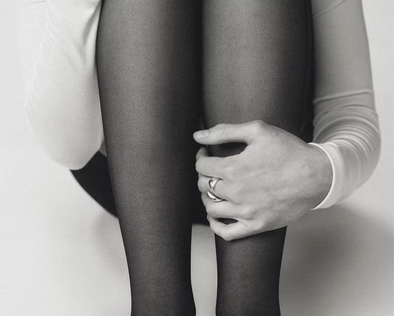 Black and white close-up of a person wearing sheer black tights, with one hand resting on the knee. The person is wearing a white long-sleeve top and a ring on one finger.