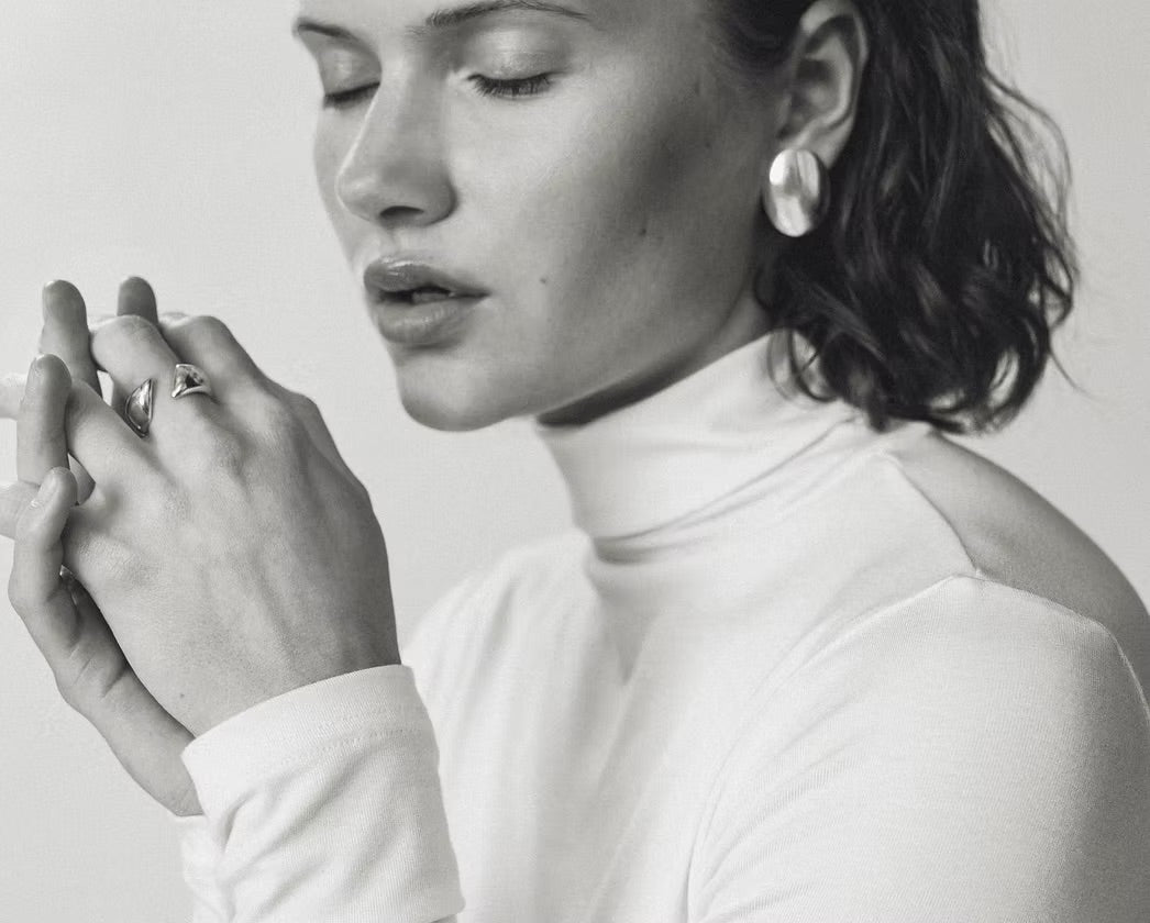 A black and white close-up portrait of a woman wearing a white turtleneck top, a large round earring, and rings on her fingers, set against a plain light background.