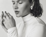 A black and white close-up portrait of a woman wearing a white turtleneck top, a large round earring, and rings on her fingers, set against a plain light background.
