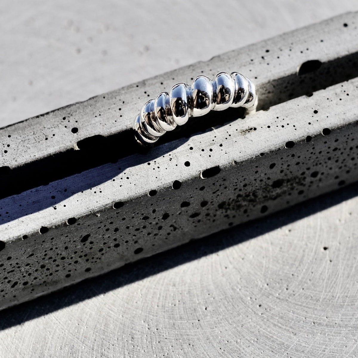 A silver metallic ring with a textured, coiled or segmented design, resting on a speckled gray concrete surface.