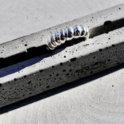 A silver metallic ring with a textured, coiled or segmented design, resting on a speckled gray concrete surface.