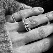 A close-up black and white photograph of a person's hand wearing a textured, multi-link ring on one finger, with a simple band ring on another finger, set against a dark knit sweater and dark fabric background.