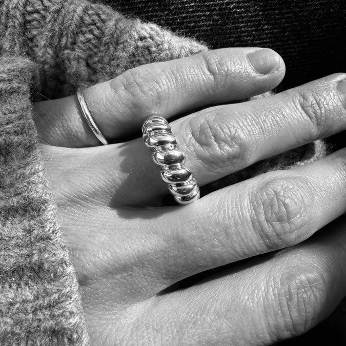 A close-up black and white photograph of a person's hand wearing a textured, multi-link ring on one finger, with a simple band ring on another finger, set against a dark knit sweater and dark fabric background.