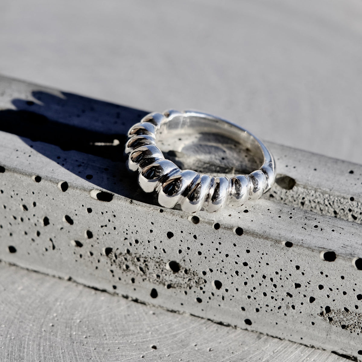 A close-up of a textured silver ring with a ridged, flower-like band, resting on a speckled gray stone surface.
