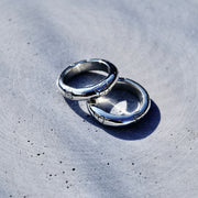 Two polished silver rings with engraved patterns, resting on a textured gray concrete surface under bright natural sunlight.