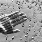 A close-up black and white photograph of a person's hand resting over wet sand covered with small pebbles, wearing a ribbed knit sweater cuff and several rings.