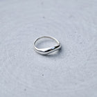 A polished silver ring with a smooth, slightly wavy, teardrop-shaped band, resting on a textured gray surface.