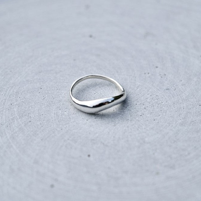 A polished silver ring with a smooth, slightly wavy, teardrop-shaped band, resting on a textured gray surface.