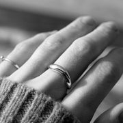 A close-up black and white photograph of a person's hand wearing a polished silver ring with a curved, organic shape. A second simple band ring is visible on the middle finger, and a textured knit sweater cuff is visible at the bottom left.