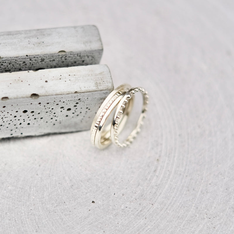 Four silver rings in different textures, displayed on a light gray stone surface next to two textured gray stone bars.