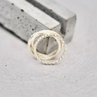Two interlocking silver rings, one with a textured beaded outer band and the other smooth, resting on a light gray textured surface next to a gray rectangular block.