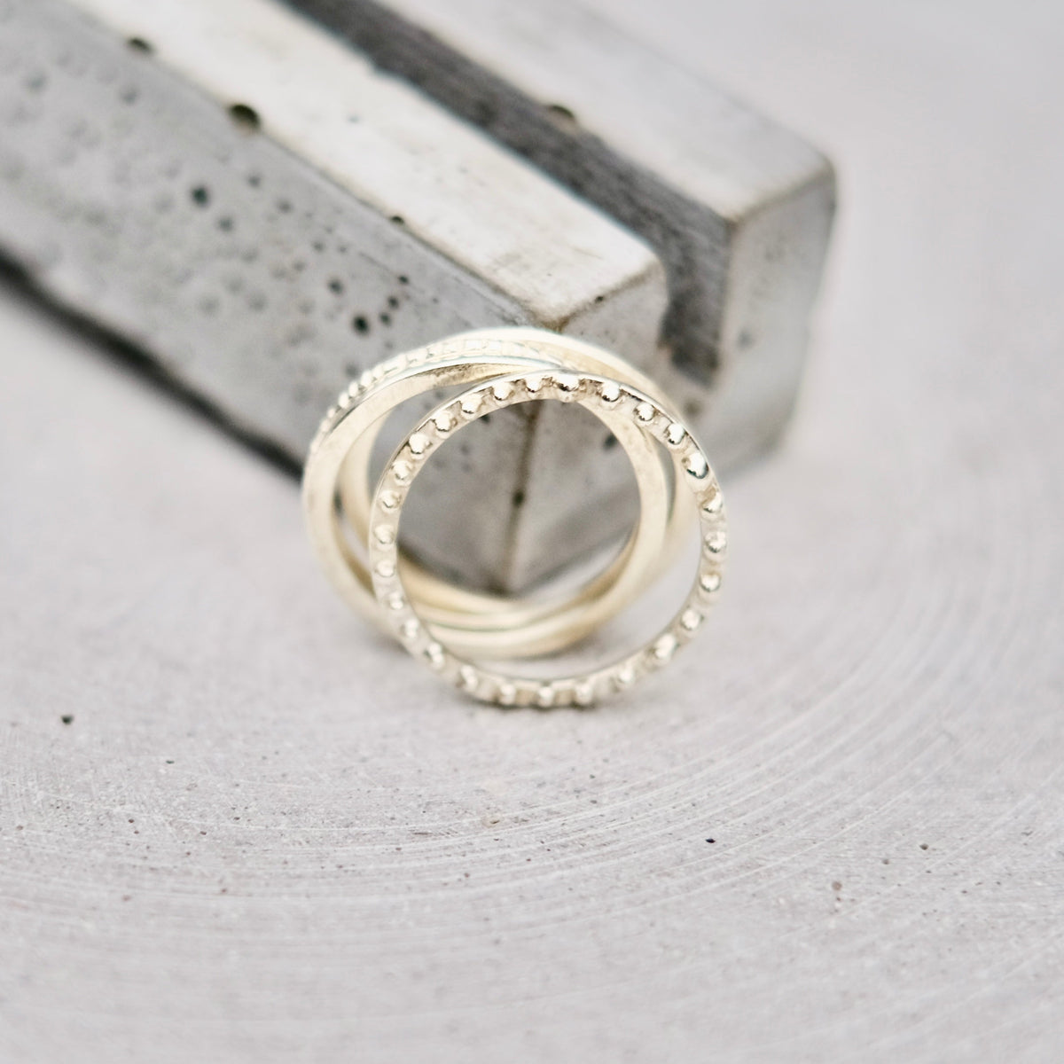 Two interlocking silver rings, one with a textured beaded outer band and the other smooth, resting on a light gray textured surface next to a gray rectangular block.