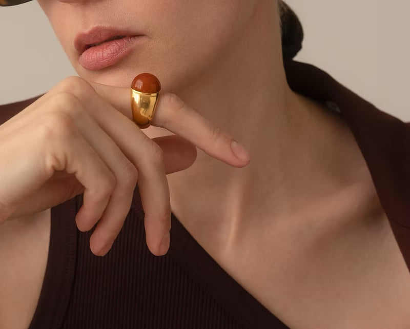 A close-up lifestyle shot of a model wearing a chunky gold-plated silver ring with a polished, rounded reddish-brown amber on her finger.