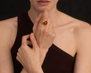 A close-up lifestyle shot of a model wearing a gold signet ring with a polished round amber-brown cabochon stone on her finger, wearing a dark brown ribbed one-shoulder top against a dark background.