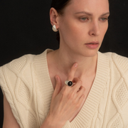 A close-up of a woman wearing a silver-toned signet ring with a black amber at the center, paired with a cream cable-knit sweater against a dark background.