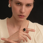 A close-up of a model wearing a chunky silver ring with a black amber center, paired with a cream cable-knit sweater against a solid black background.