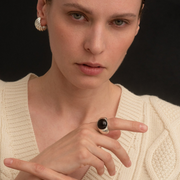 A close-up of a model wearing a chunky silver ring with a black amber center, paired with a cream cable-knit sweater against a solid black background.