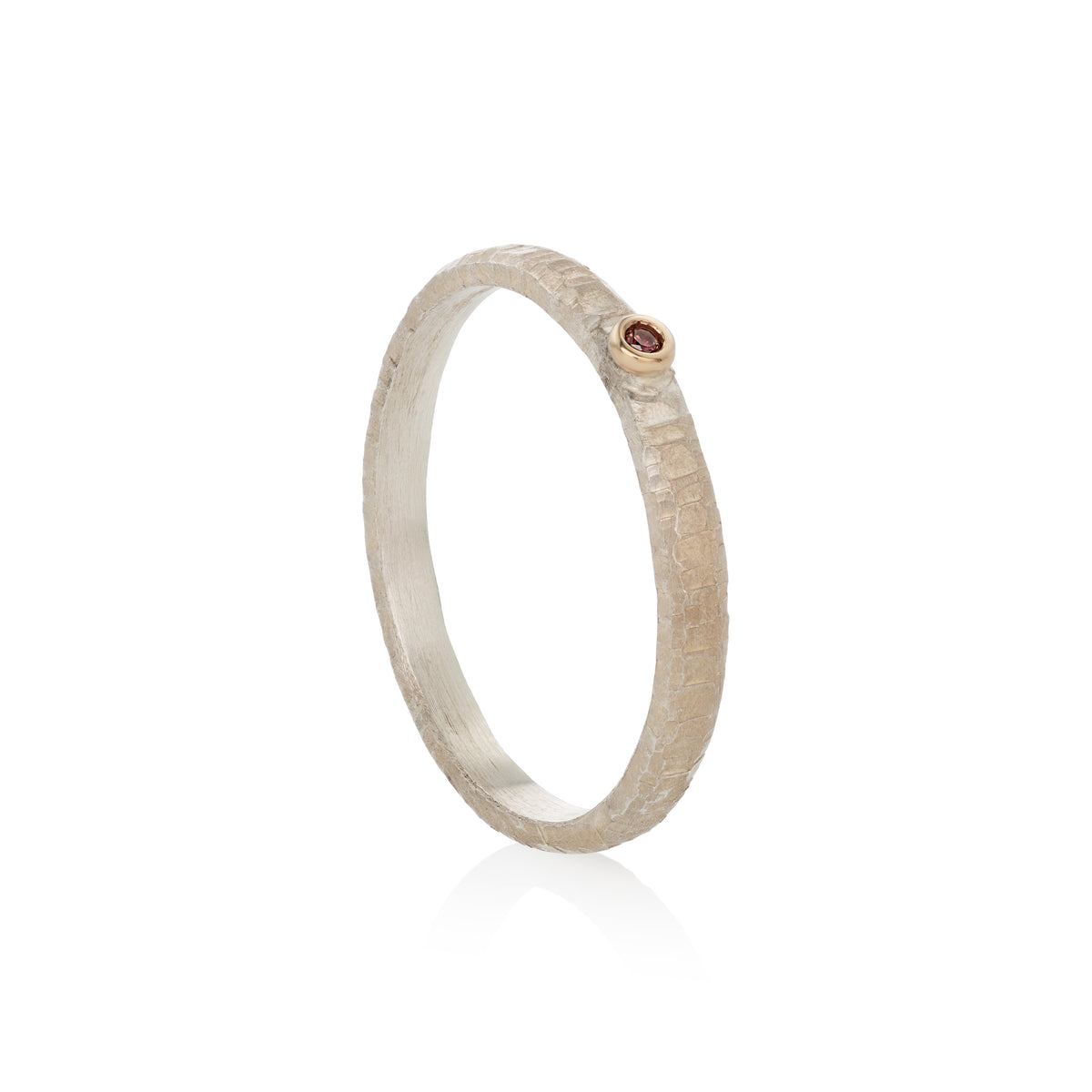A textured silver ring with a small round gold bezel holding a tiny red diamond, shown on a clean white background with a soft reflection.
