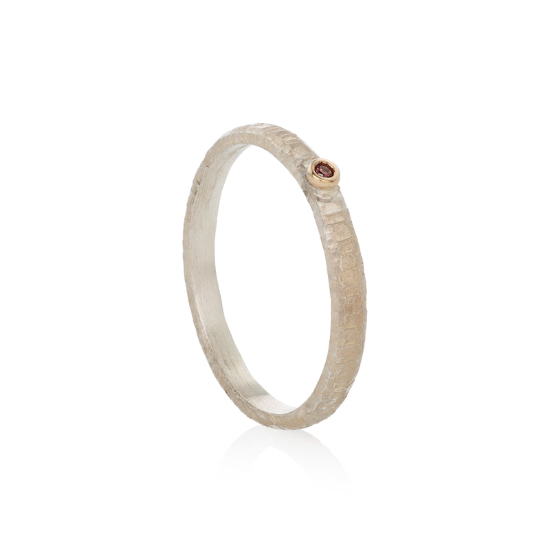A textured silver ring with a small round gold bezel holding a tiny red diamond, shown on a clean white background with a soft reflection.