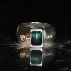 A silver ring featuring a rectangular green iridescent gemstone and two small moissnaites on the band, photographed on a plain light background.