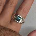 A silver ring featuring a rectangular green iridescent gemstone and two small moissnaites on the band, photographed on a plain light background.
