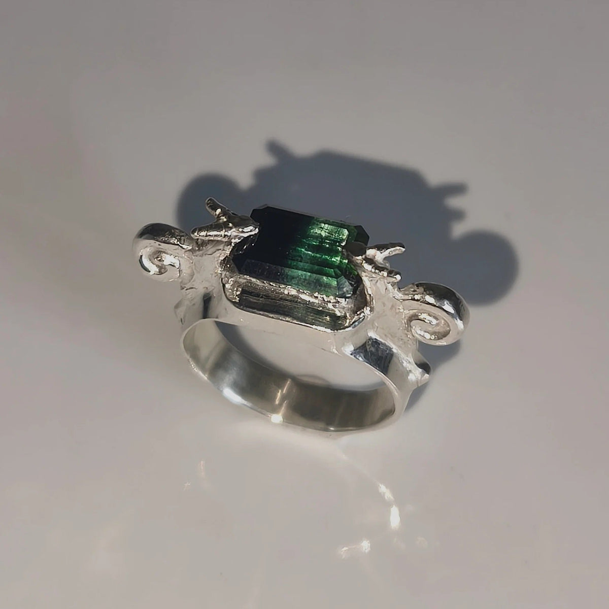 A silver ring featuring a rectangular green iridescent gemstone, photographed on a light gray reflective surface with a soft shadow.