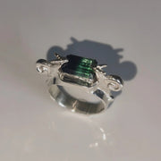 A silver ring featuring a rectangular green iridescent gemstone, photographed on a light gray reflective surface with a soft shadow.