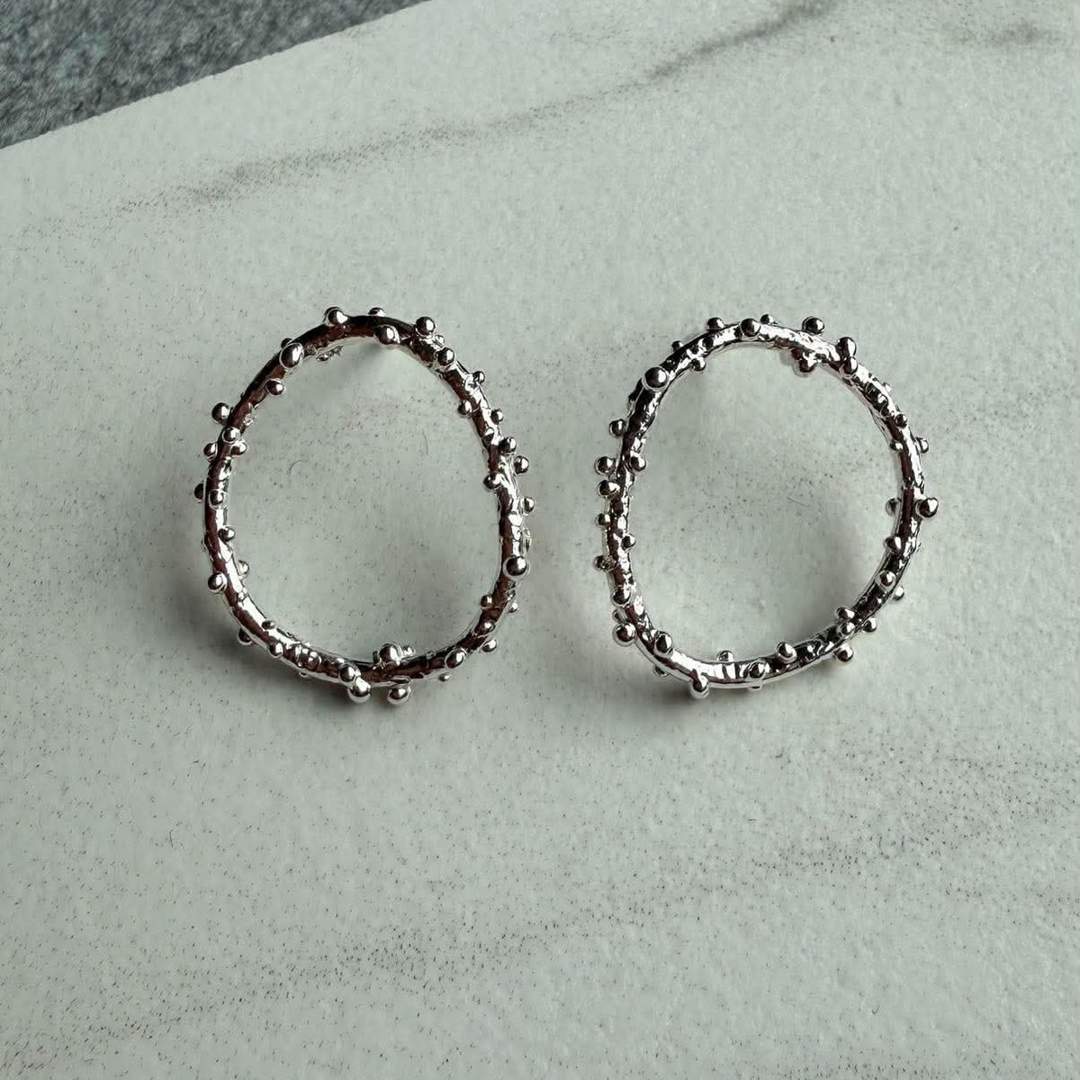 A pair of silver open-circle earrings featuring a textured, beaded surface,  displayed on a light gray marble surface.