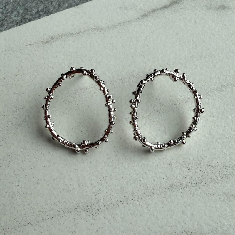 A pair of silver open-circle earrings featuring a textured, beaded surface,  displayed on a light gray marble surface.