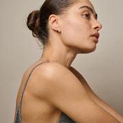 A close-up side profile of a woman wearing a small silver crescent moon stud earring with a clear gemstone, set against a neutral beige background.