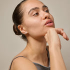 A close-up portrait of a woman with her hair in a bun, wearing a pair of small silver stud earrings, set against a plain beige background.
