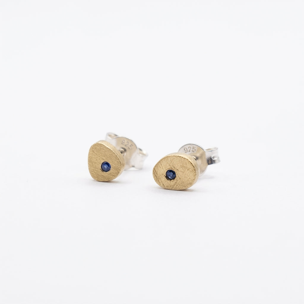 A pair of textured gold and silver stud earrings, each featuring a small round dark blue gemstone, shown on a clean white background.