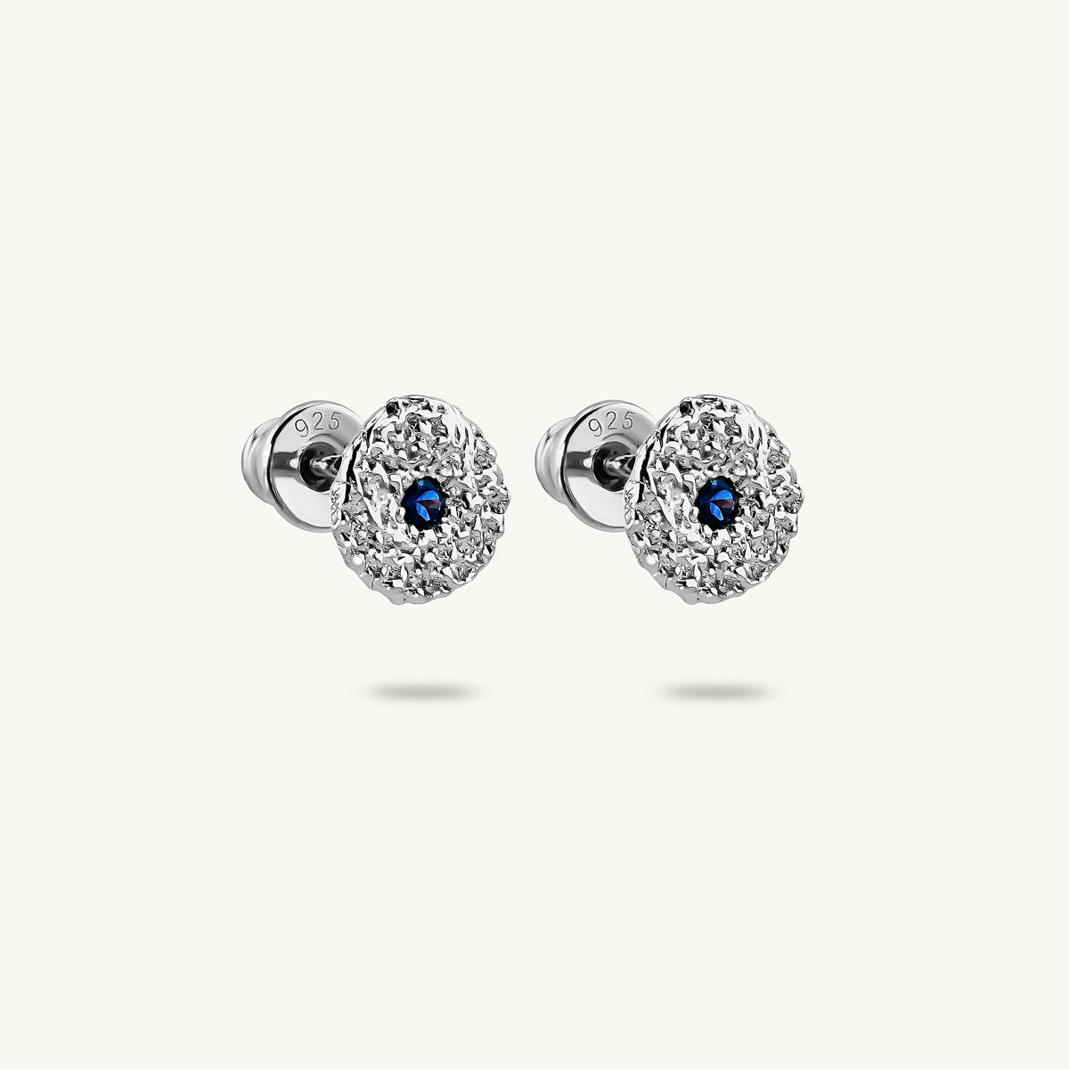 A pair of silver stud earrings with a textured, flower-like circular face and a small round blue sapphires in the center, shown on a light off-white background.