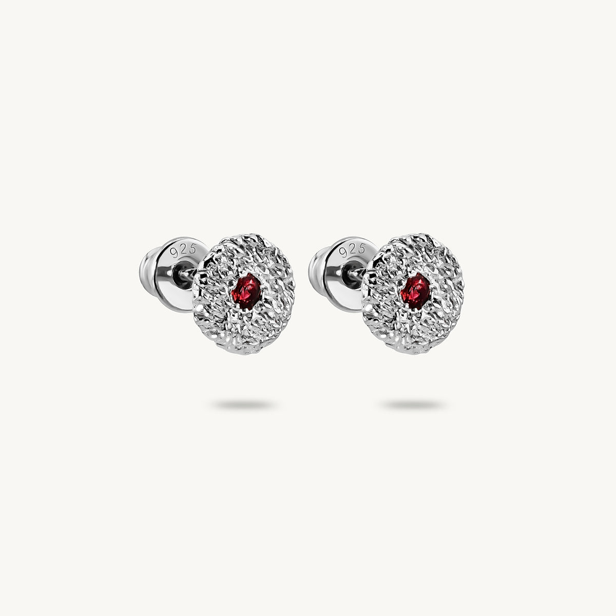 A pair of silver stud earrings with a textured, flower-like circular face and a small round red sapphires in the center, shown on a light off-white background.