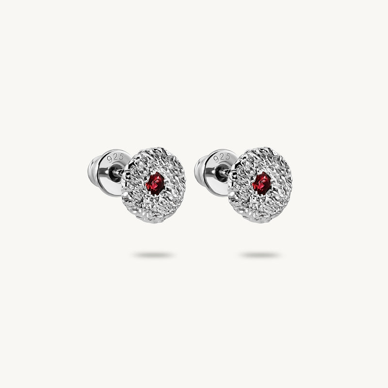 A pair of silver stud earrings with a textured, flower-like circular face and a small round red sapphires in the center, shown on a light off-white background.