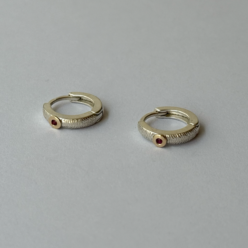 A pair of silver hoop earrings with small rubies attached to them, set against a light background.