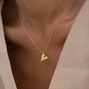 A gold plated silver necklace with a heart-shaped pendant, worn by a person with a light skin tone, presented in a close-up shot focusing on the necklace.