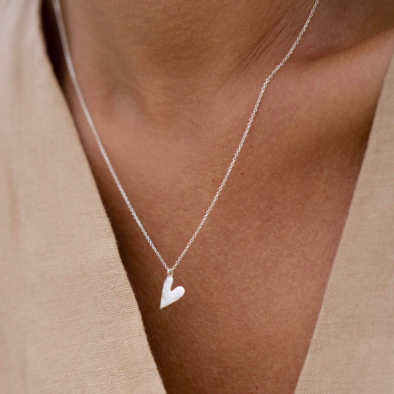 A heart-shaped silver necklace on a person's neck, presented as a gift in a designer-stamped box.