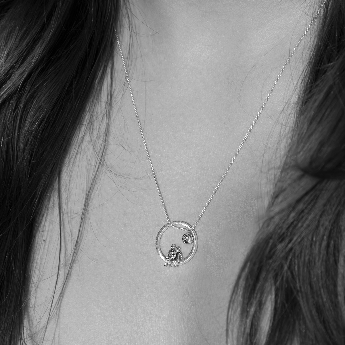 Necklace with a circular pendant featuring a small human figurines and diamond, worn by a person with long hair.