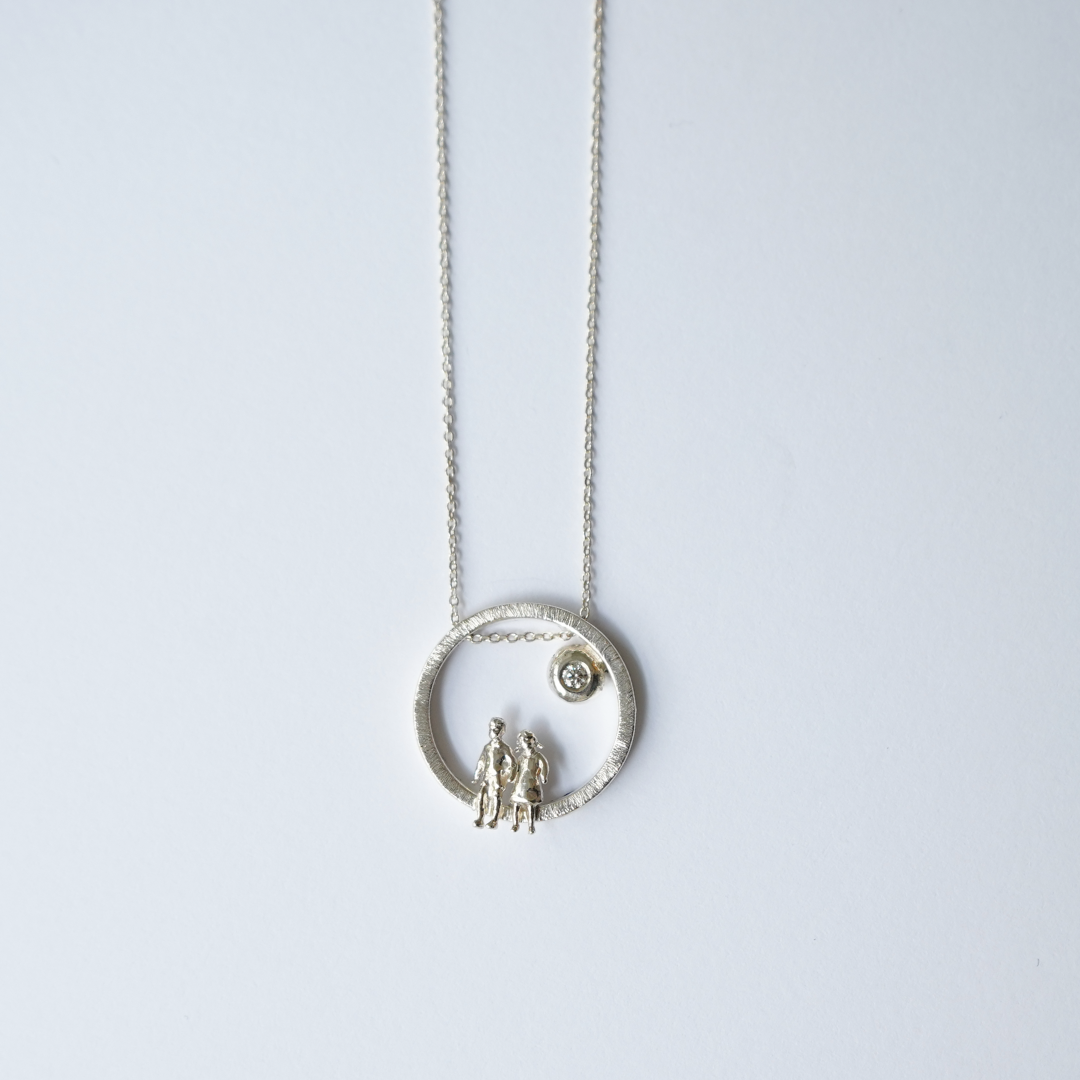 A silver necklace featuring a textured circular pendant with two small human figures and a tiny sun detail, suspended from a delicate silver chain against a light gray background.