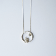 A silver necklace featuring a textured circular pendant with two small human figures and a tiny sun detail, suspended from a delicate silver chain against a light gray background.