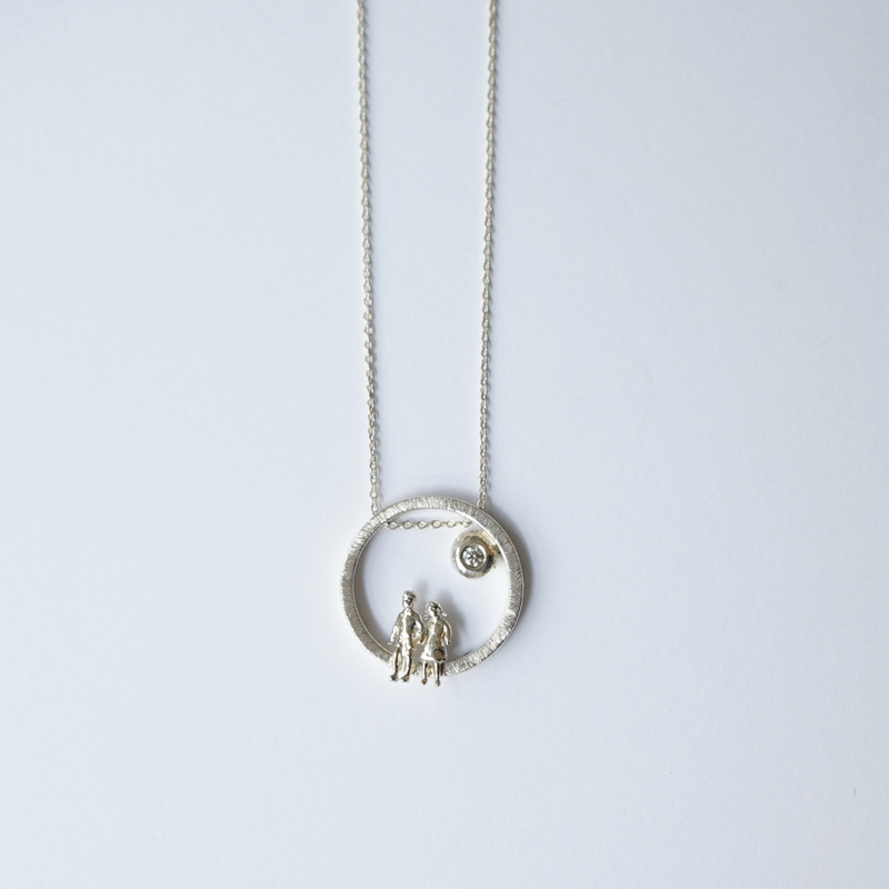 A silver necklace featuring a textured circular pendant with two small human figures and a tiny sun detail, suspended from a delicate silver chain against a light gray background.