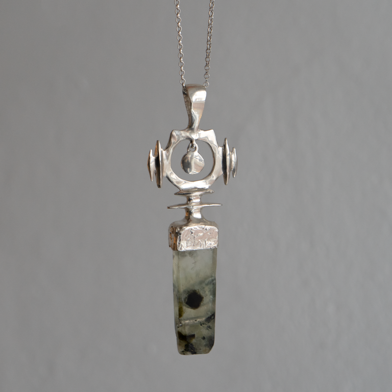 A handmade silver pendant necklace featuring a polished greenish-gray stone with dark inclusions, suspended from a textured silver bail on a fine chain, set against a neutral gray background.