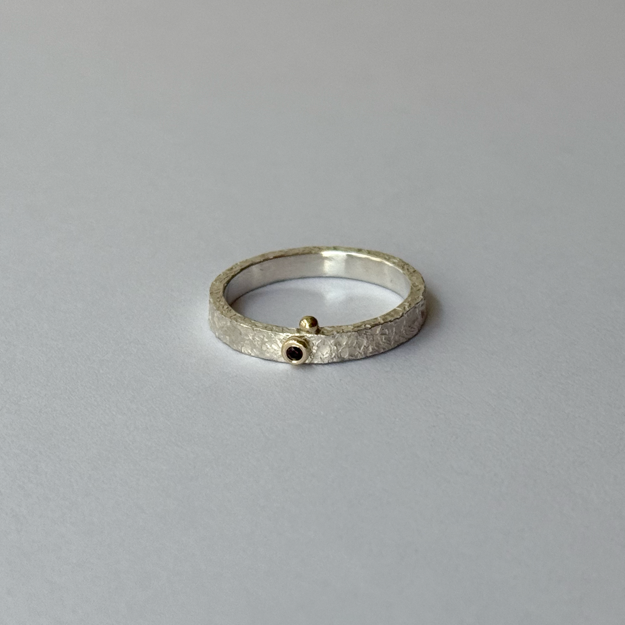 A silver ring with a red garnet in the center, decorated with gold bubbles, placed on a light background.
